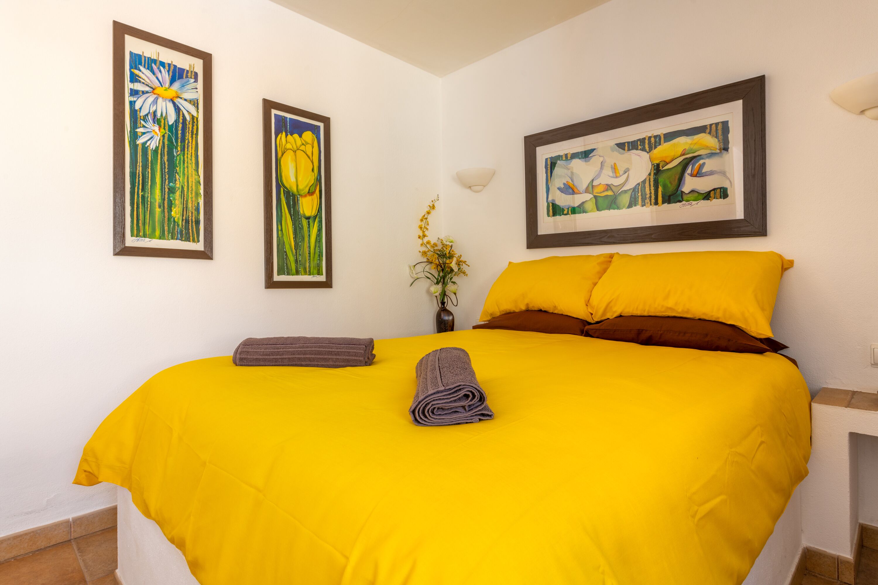 Une chambre d’h ôtel avec un grand lit, une literie jaune et deux tableaux représentant des motifs floraux, encadrés et accrochés au mur.