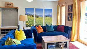 Een woonkamer met een blauwe zithoek, een houten salontafel en uitzicht op een berglandschap door grote ramen.