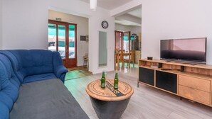 Ein modernes Wohnzimmer mit einem blauen Sofa, einem Couchtisch mit Bierflaschen, einem Fernseher auf einem Holzständer und einer Küche im Hintergrund.