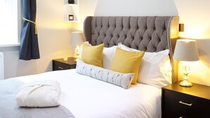 A neatly made bed with a grey headboard, white bedding, and a few pillows in yellow and white. There is a nightstand with a lamp and a small shelf above it. - Avari Apartments - Winchester Collection (London)