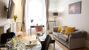 A modern living room with a grey sofa, a glass coffee table, and a dining table set with wine glasses and plates. - Avari Apartments - Winchester Collection (London)