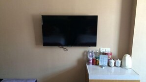 Een kamer met een wandgemonteerde flatscreen-tv, een klein tafeltje met diverse spullen, waaronder flessen, een waterkoker en bekers.