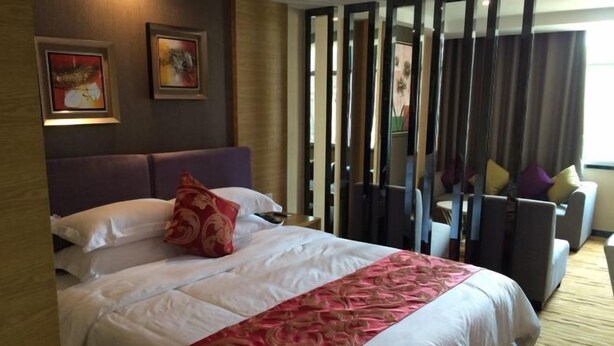 Hotelkamer met een groot bed, hoofdeinde en zicht door een scheidingswand op een zithoek met een bank en stoelen.