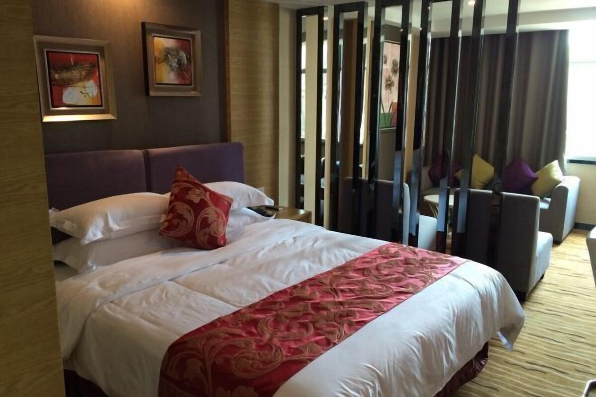 Habitación de hotel con una cama grande, cabecera y vistas a una zona de estar con sofá y sillones, separadas por un biombo.