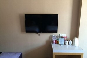 Una habitación con una televisión de pantalla plana montada en la pared, una mesita con diversos objetos, incluyendo botellas y un hervidor, y una silla morada.