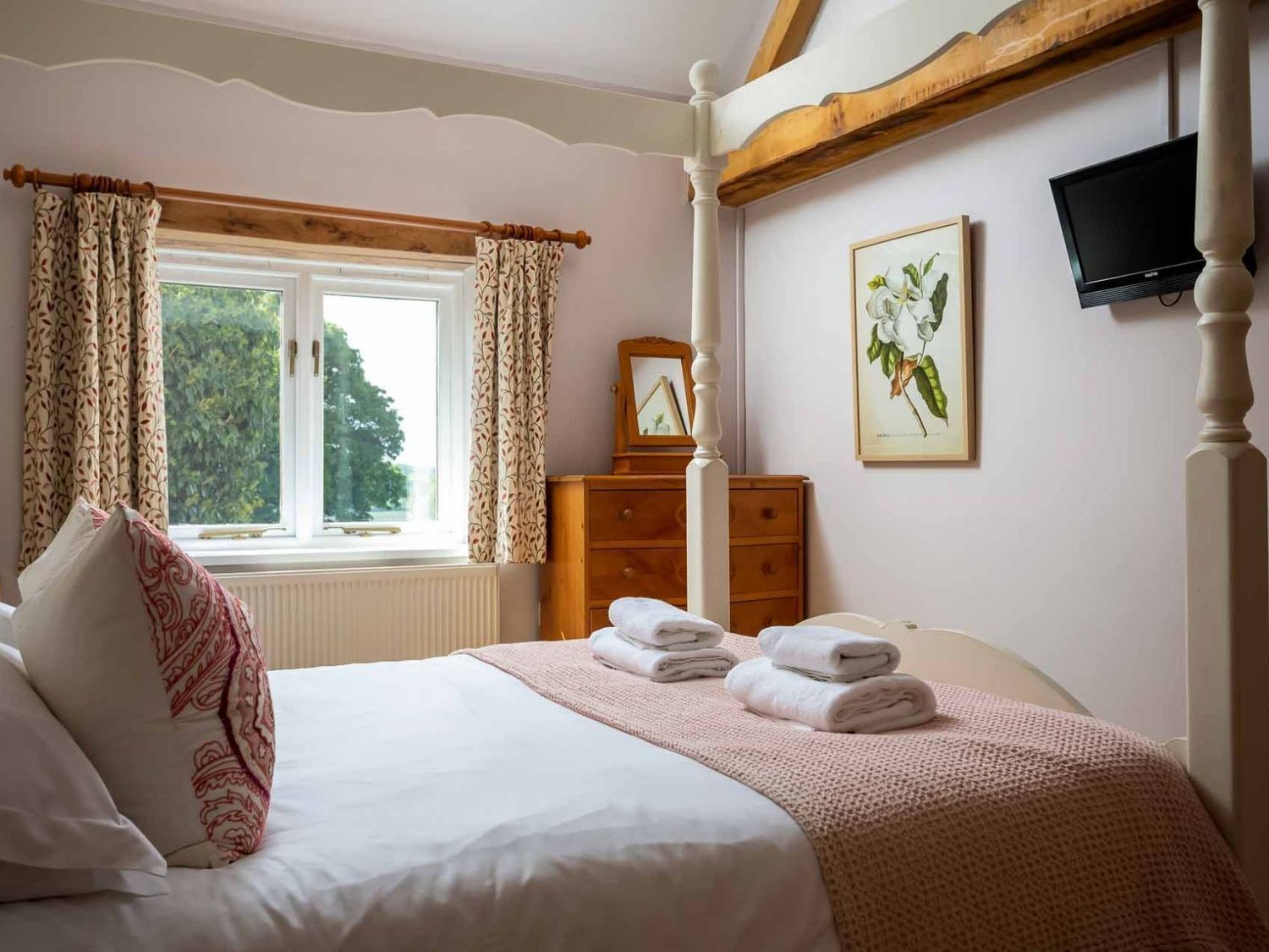 A bedroom with a bed, a dresser, a window with curtains, a painting, and a television on the wall.