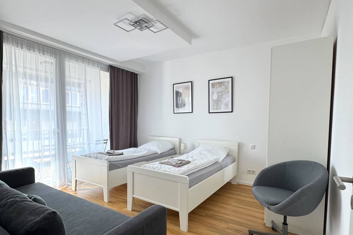 A modern hotel room with two single beds, a grey sofa, a desk, and a chair.