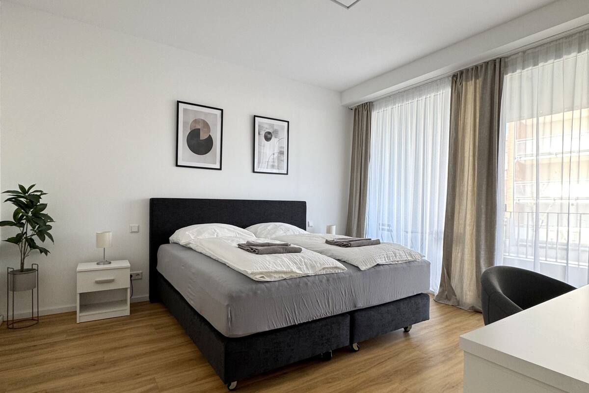 A modern bedroom with a large bed, two framed artworks, a bedside table with a lamp, a potted plant, and a view of the city through sheer curtains.