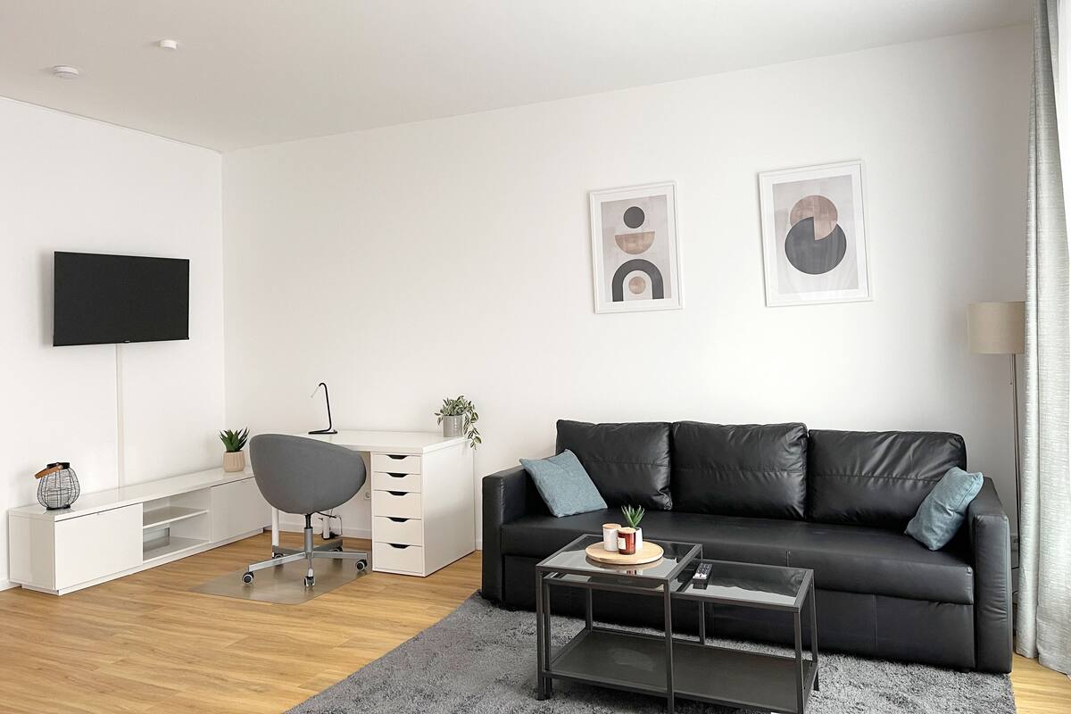 A modern living room with a black leather sofa, a glass coffee table, a white desk with a chair, a flat-screen TV mounted on the wall, and abstract art pieces.