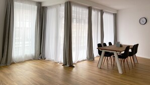 Una habitación amplia con una mesa y sillas de madera, ventanas grandes con cortinas translúcidas y un reloj de pared.
