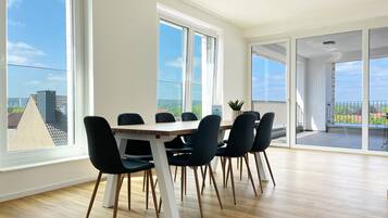 A spacious room with a wooden floor, a large dining table with chairs, and large windows offering a view of the cityscape.