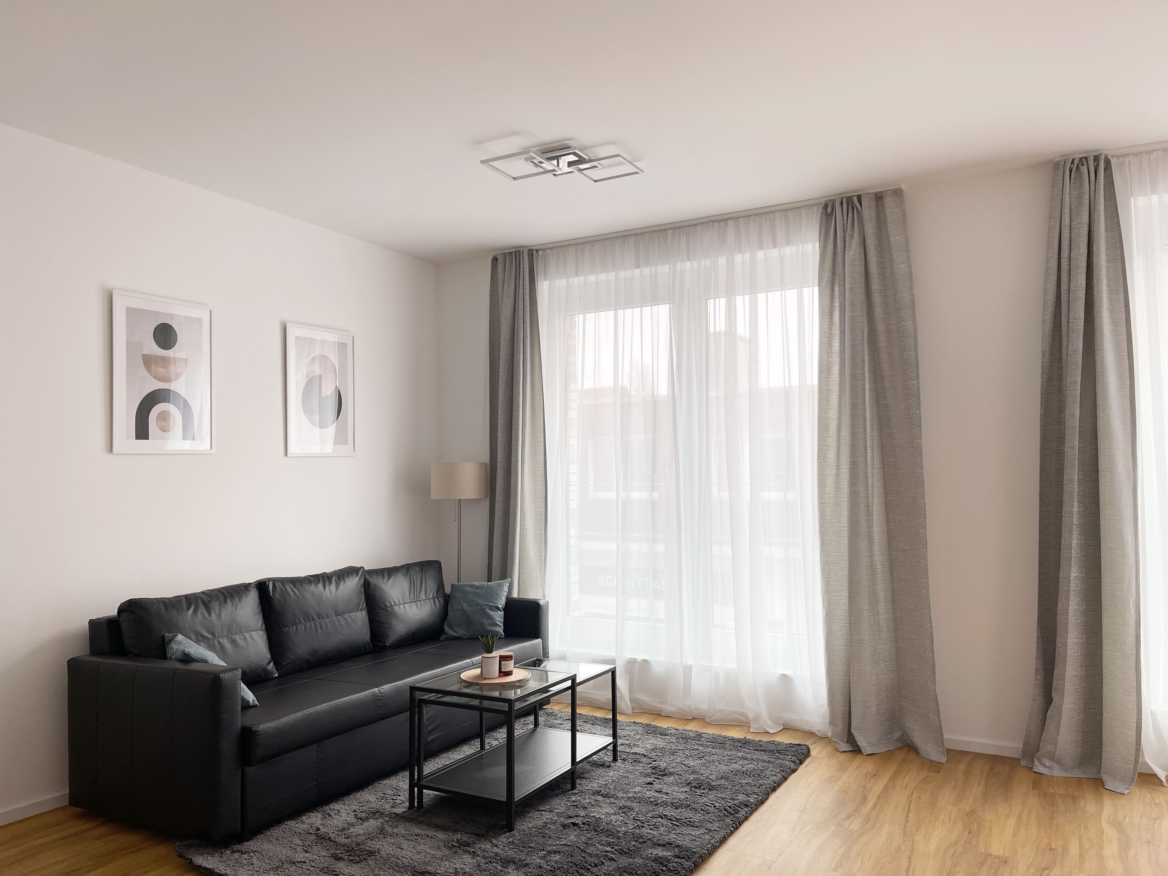 Een moderne woonkamer met een zwarte leren bank, een glazen salontafel en een grijs vloerkleed. Twee ingelijste abstracte kunstwerken hangen aan de muur, een vloerlamp en grote grijze gordijnen bedekken de ramen.