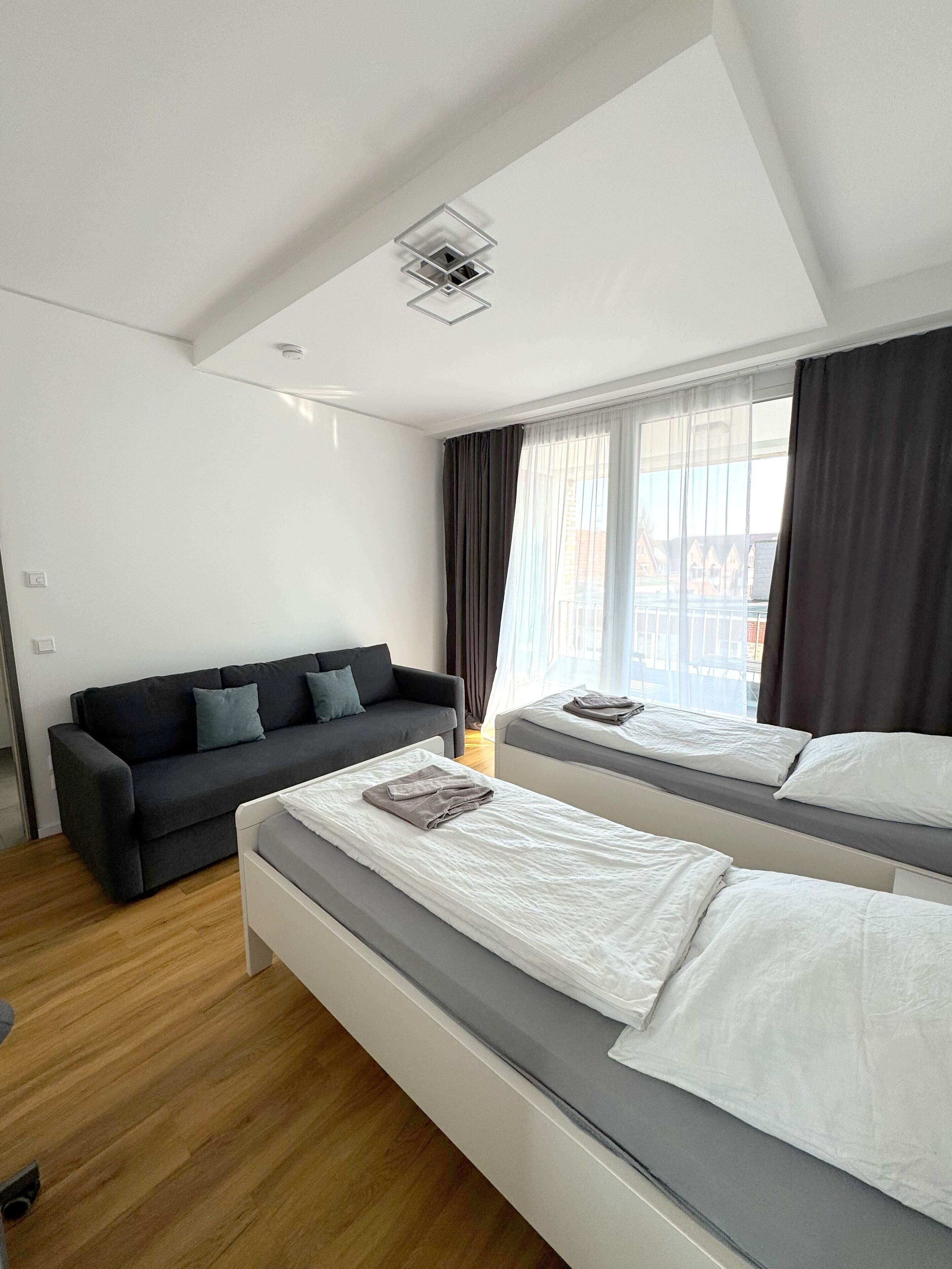 A modern hotel room with two single beds, a dark-colored sofa, and a large window with curtains.