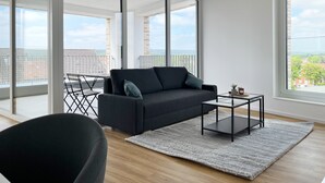 Un salón moderno con un sofá gris oscuro, una mesa de centro de cristal y una alfombra gris. La habitación tiene ventanas amplias que ofrecen vistas de la ciudad y un balcón con barandilla de metal.
