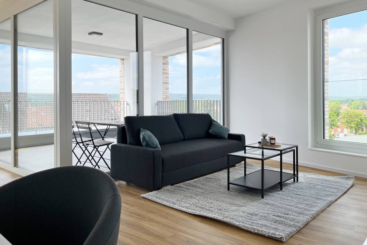 A modern living room with a dark grey sofa, a glass coffee table, and a grey rug. The room has large windows offering a view of the cityscape and a balcony with a metal railing.
