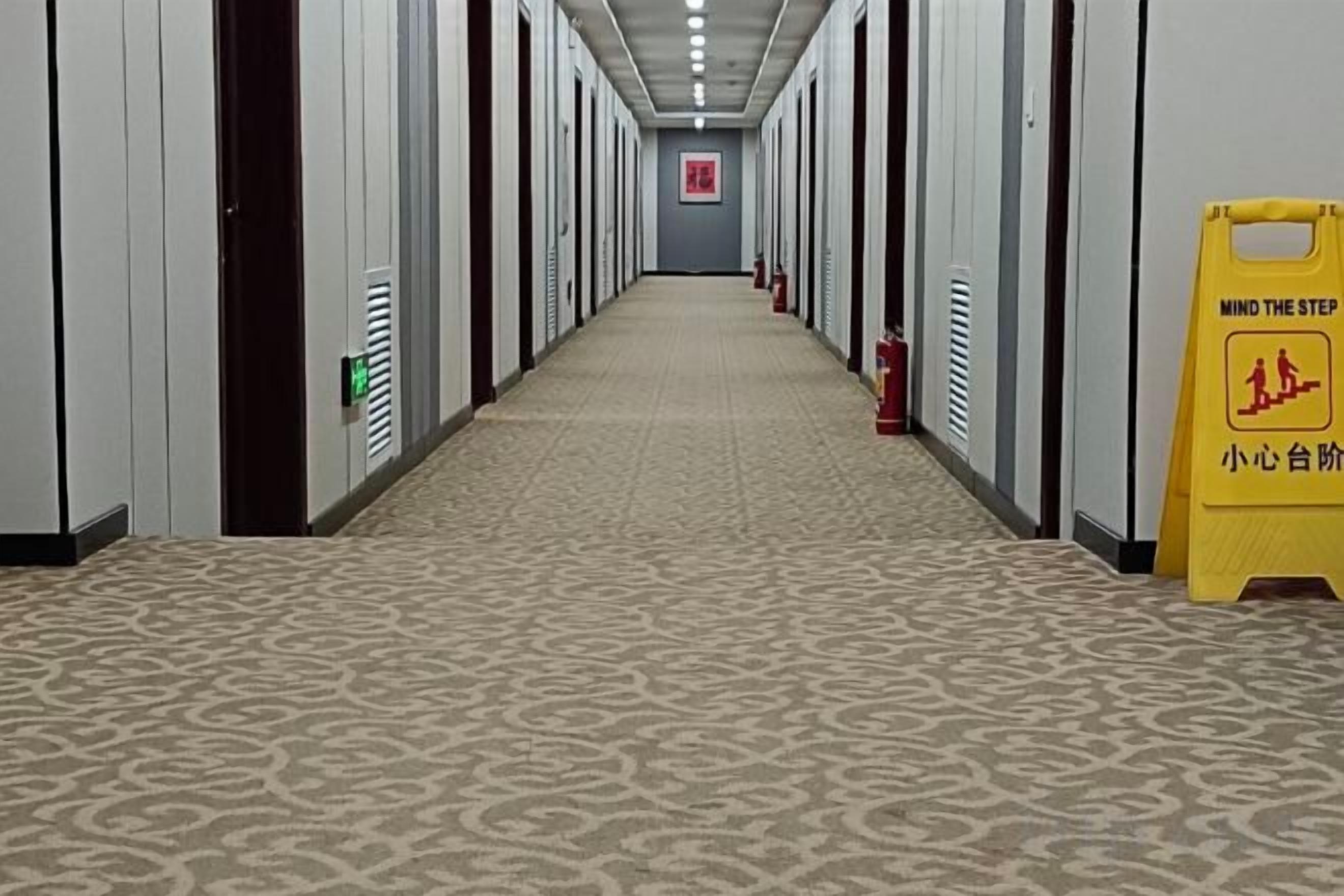 Un pasillo de hotel con alfombra estampada, puertas a ambos lados y un letrero amarillo con texto.