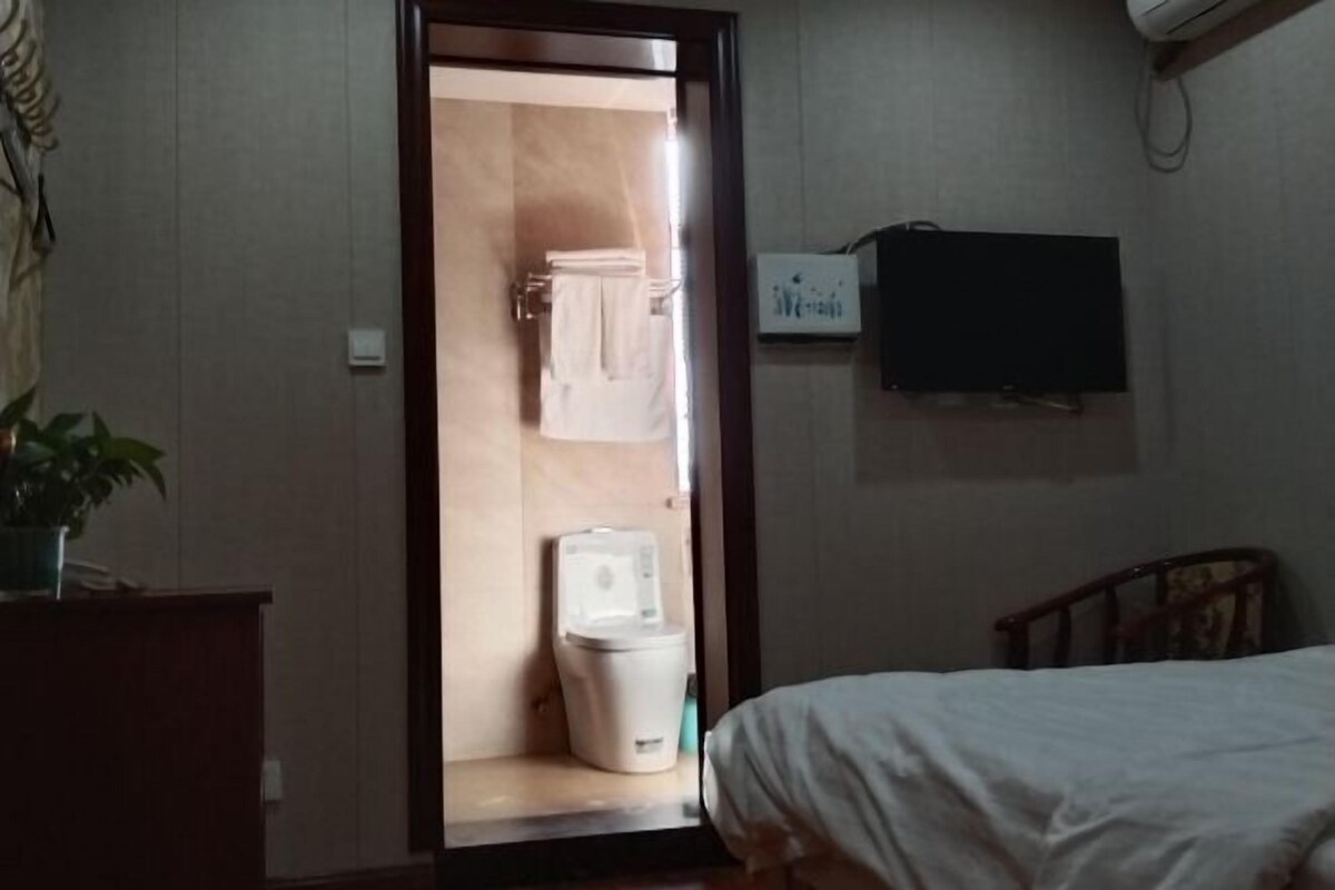 A hotel room with a bed, a TV mounted on the wall, a wooden headboard, and a small desk with a plant.