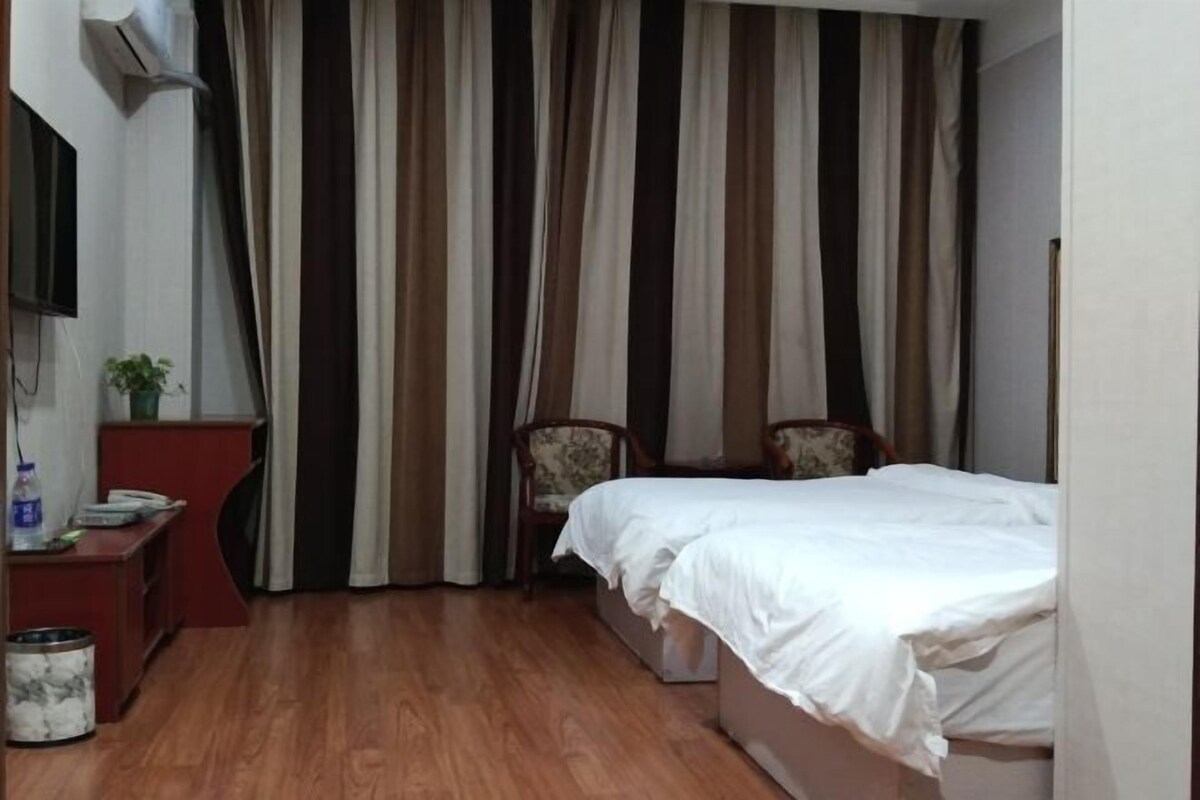 A hotel room with a wooden floor, a bed with white bedding, a desk with a telephone and a plant, and a chair.