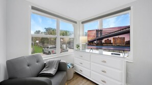 Quarto com sofá cinzento, cómoda branca e grande janela com pintura de uma ponte.