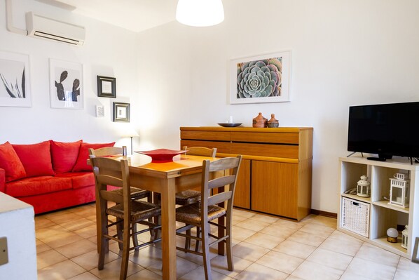 A living room with a red sofa, wooden dining table, chairs, a TV, and a kitchen area with cabinets and a countertop.