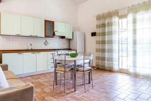 Une cuisine avec des meubles blancs, une table à manger avec des chaises, un saladier vert et une fenêtre avec des rideaux.