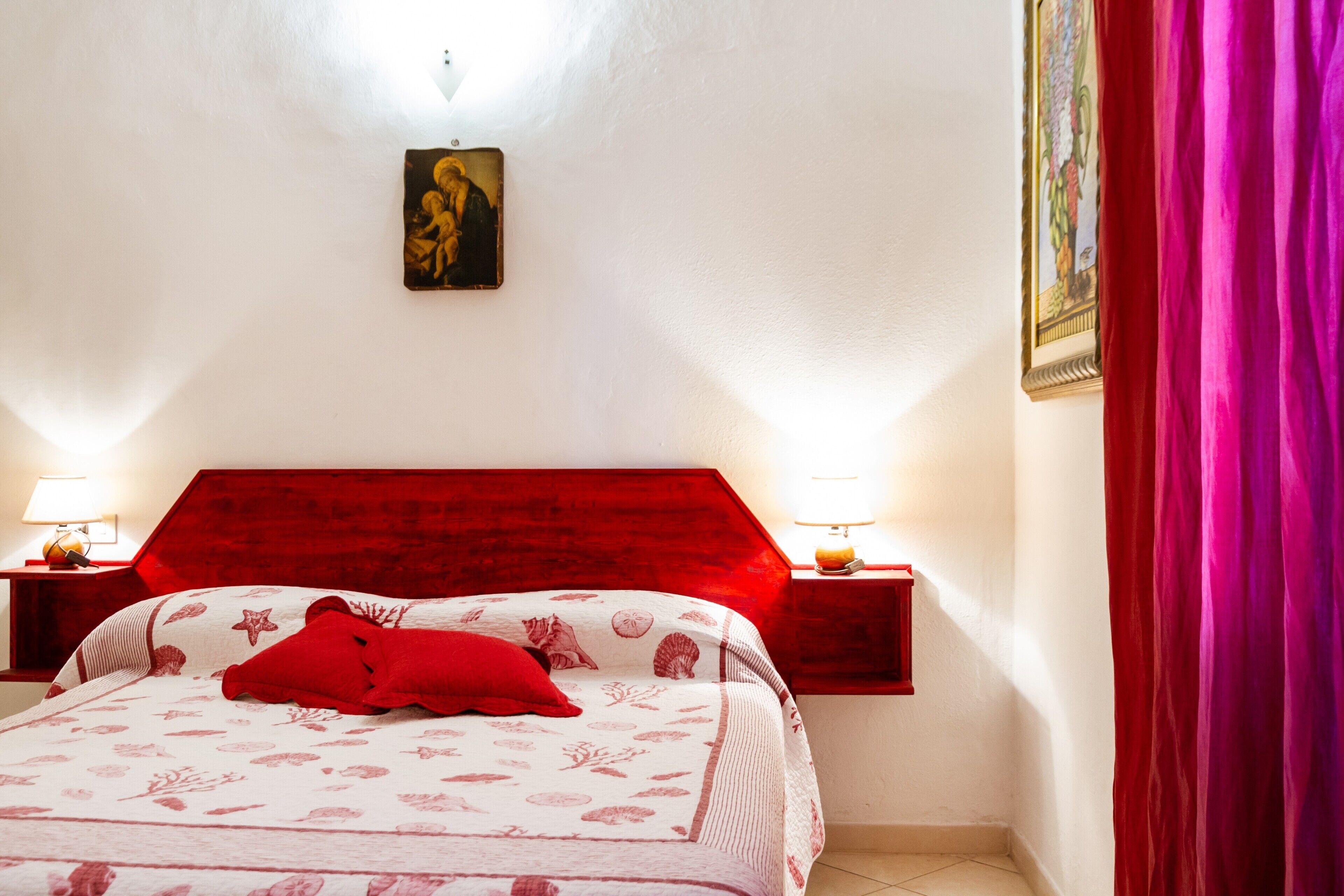 Un dormitorio con cabecera roja, dos lámparas de noche, un almohá de color rojo y un cuadro enmarcado en la pared.