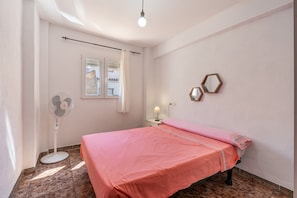 A bedroom with a bed, a standing fan, a window with curtains, and a wall-mounted mirror.