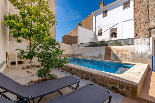 A poolside area with lounge chairs, a small tree, and a white building with a stone wall.