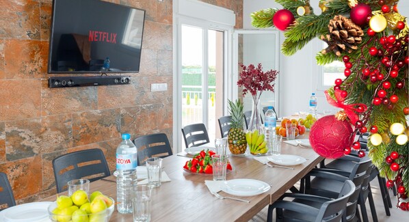 A dining area with a stone wall, a flat-screen TV, a Christmas tree, and a table set for four with fruits and beverages.