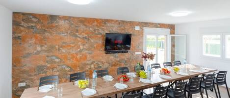 A dining area with a long table, chairs, and a TV mounted on a stone wall.