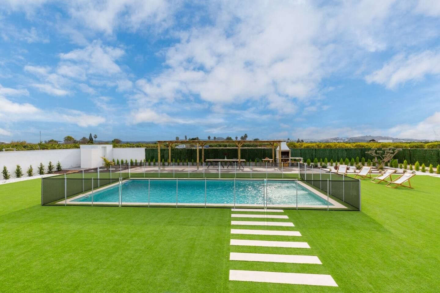 Piscina retangular com um deck ao redor, uma pérgola e uma fileira de espreguiçadeiras.