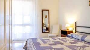 Une chambre d’hôtel avec un lit, une chaise, une table de chevet avec une lampe, un miroir et une fenêtre avec des rideaux.