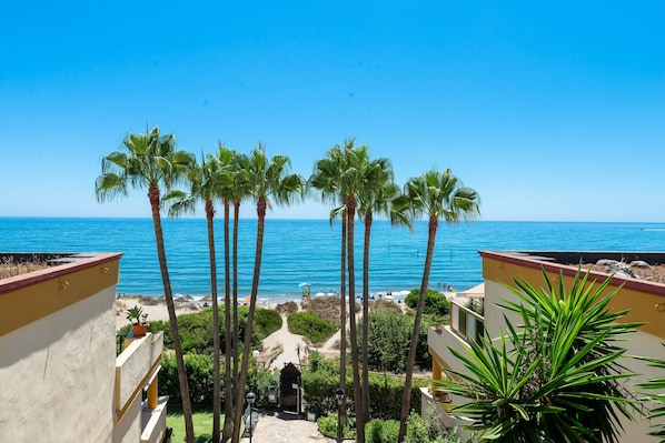 A balcony with a view of the beach and ocean, palm trees, and a building with a yellow facade.