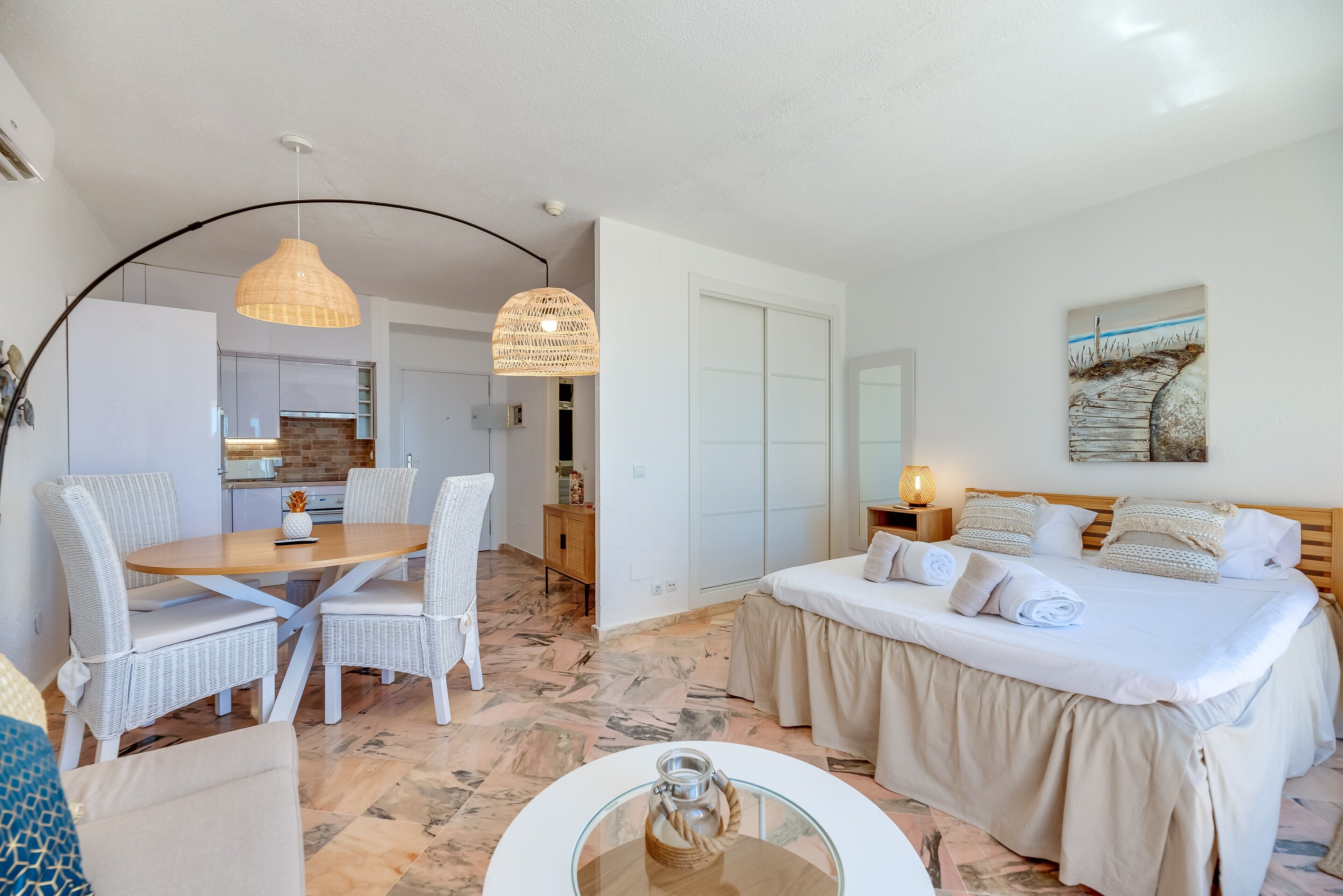 Un dormitorio con cama, mesitas de noche, una zona de comedor con sillas, una cocina al fondo y un pasillo que conduce a un armario.
