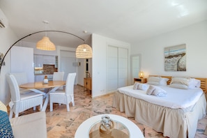 A bedroom with a bed, bedside tables, a dining area with chairs, a kitchen in the background, and a hallway leading to a closet.