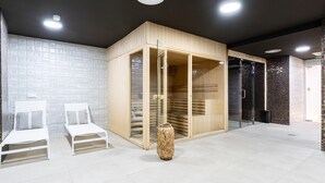 Une salle de spa moderne avec deux fauteuils blancs, un sauna en bois et un espace de douche clos par des parois de verre.
