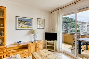 Une pièce avec un bureau en bois, un téléviseur posé sur un meuble, un tableau et un balcon donnant sur des immeubles.