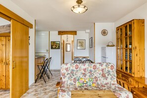Una sala de estar acogedora con un sofá floral, muebles de madera y un comedor con sillas, además de una cocina al fondo.