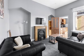A modern living room with a fireplace, a flat-screen TV, and a dining area with a kitchen visible through an open door. - Sommerset Cottages (Sunderland)