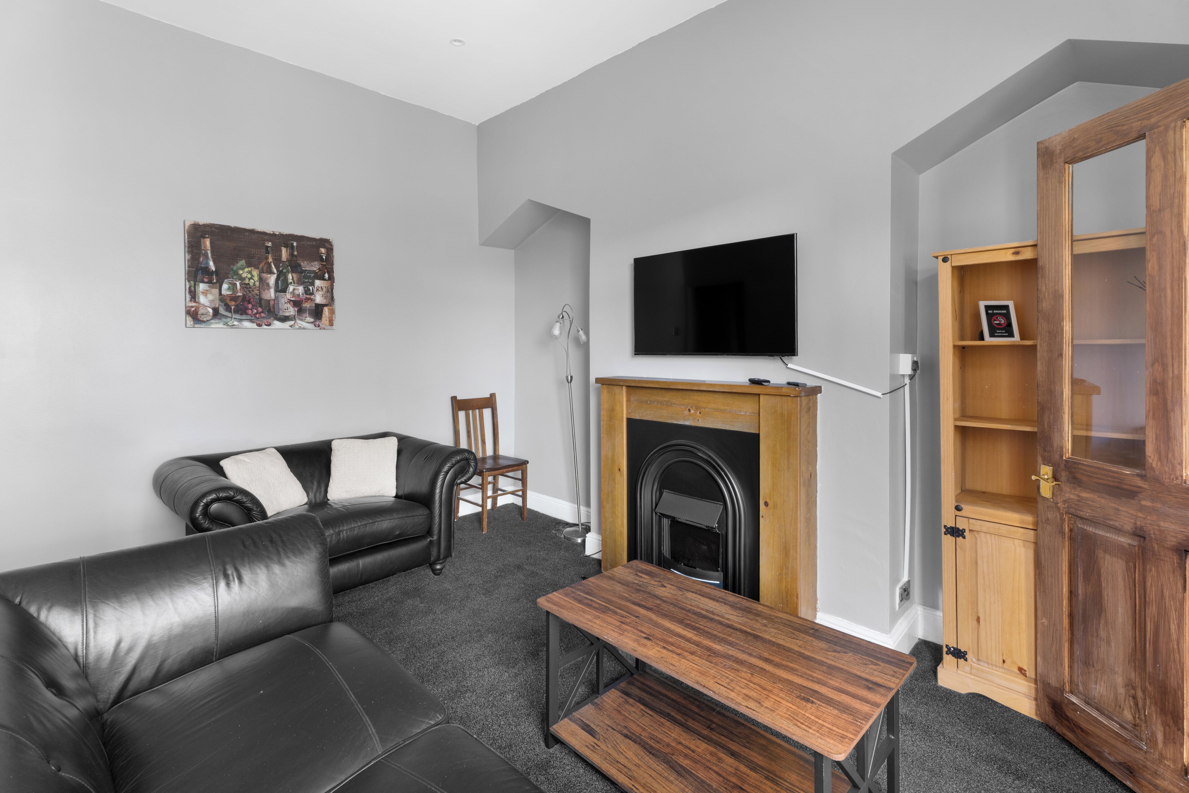 A modern living room with a black leather sofa, a wooden coffee table, a flat-screen TV mounted above a fireplace, and a painting on the wall.