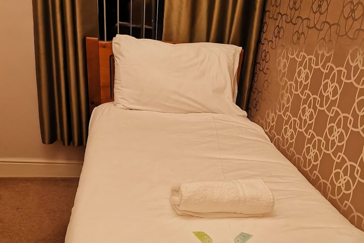 Un lit bien fait, avec un drap blanc et une serviette pliée, une tête de lit en bois et un mur décoré d’un motif.