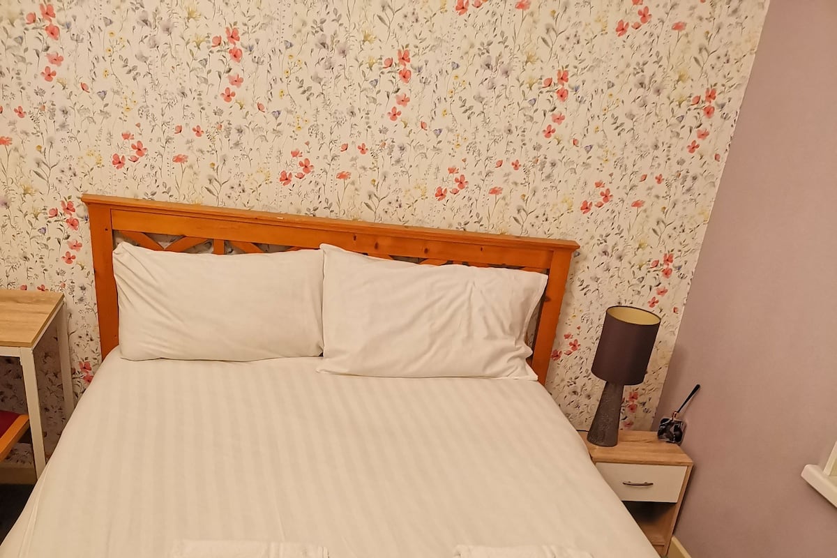 Un lit bien fait, avec du linge de lit blanc et deux serviettes pliées, une tête de lit en bois, une table de chevet avec une lampe et un papier peint à motifs floraux.