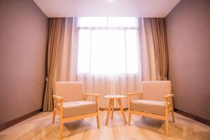 Habitació amb dos seients beixos i una taula rodona petita, una finestra gran amb cortines lleugeres i un sòl de fusta.