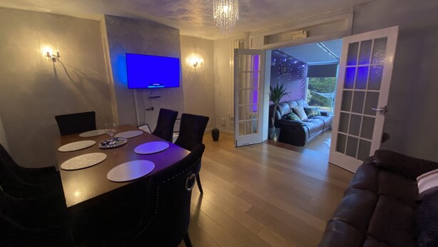 A dining area with a wooden table, chairs, and a flat-screen TV. There is a sofa and a door leading to a balcony with a view. - Impeccable 3- Bedroom House in Kirkcaldy (Kirkcaldy)