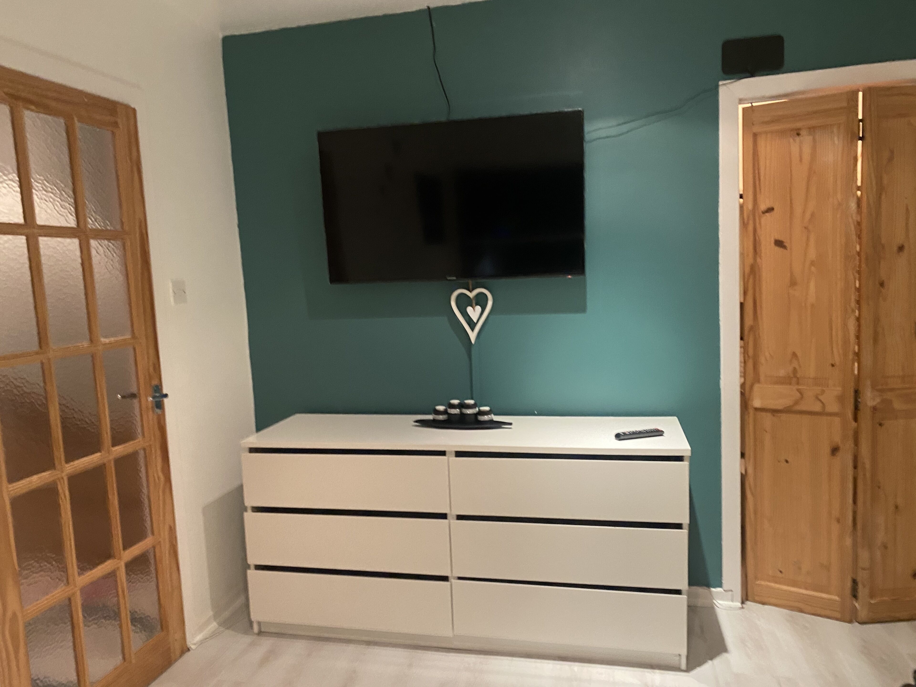A room with a teal wall, a white dresser with drawers, a flat-screen TV mounted above it, and a wooden door.