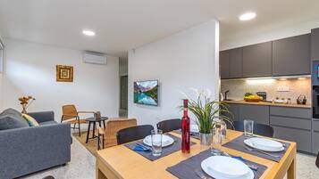 A modern living room with a dining table set for two, a grey sofa, and a kitchen with grey cabinets and a countertop.