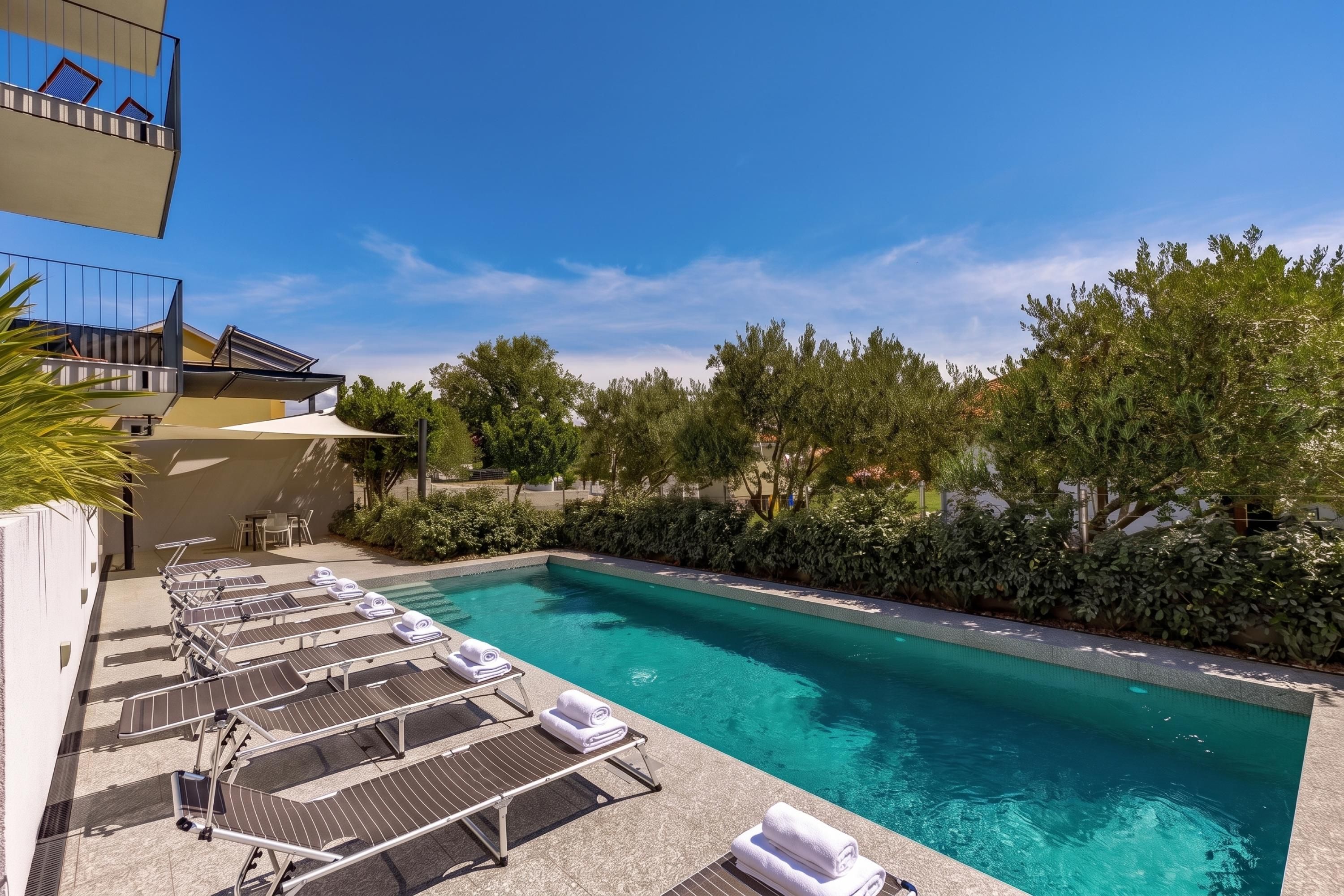 A swimming pool with lounge chairs and sun loungers, surrounded by greenery and a clear sky.