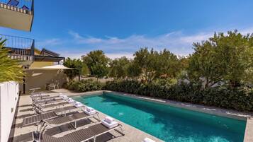 A swimming pool with lounge chairs and sun loungers, surrounded by greenery and a clear sky.