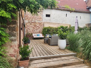 Una terraza de madera con mobiliario exterior, un muro de piedra y un edificio con techo de tejas rojas.
