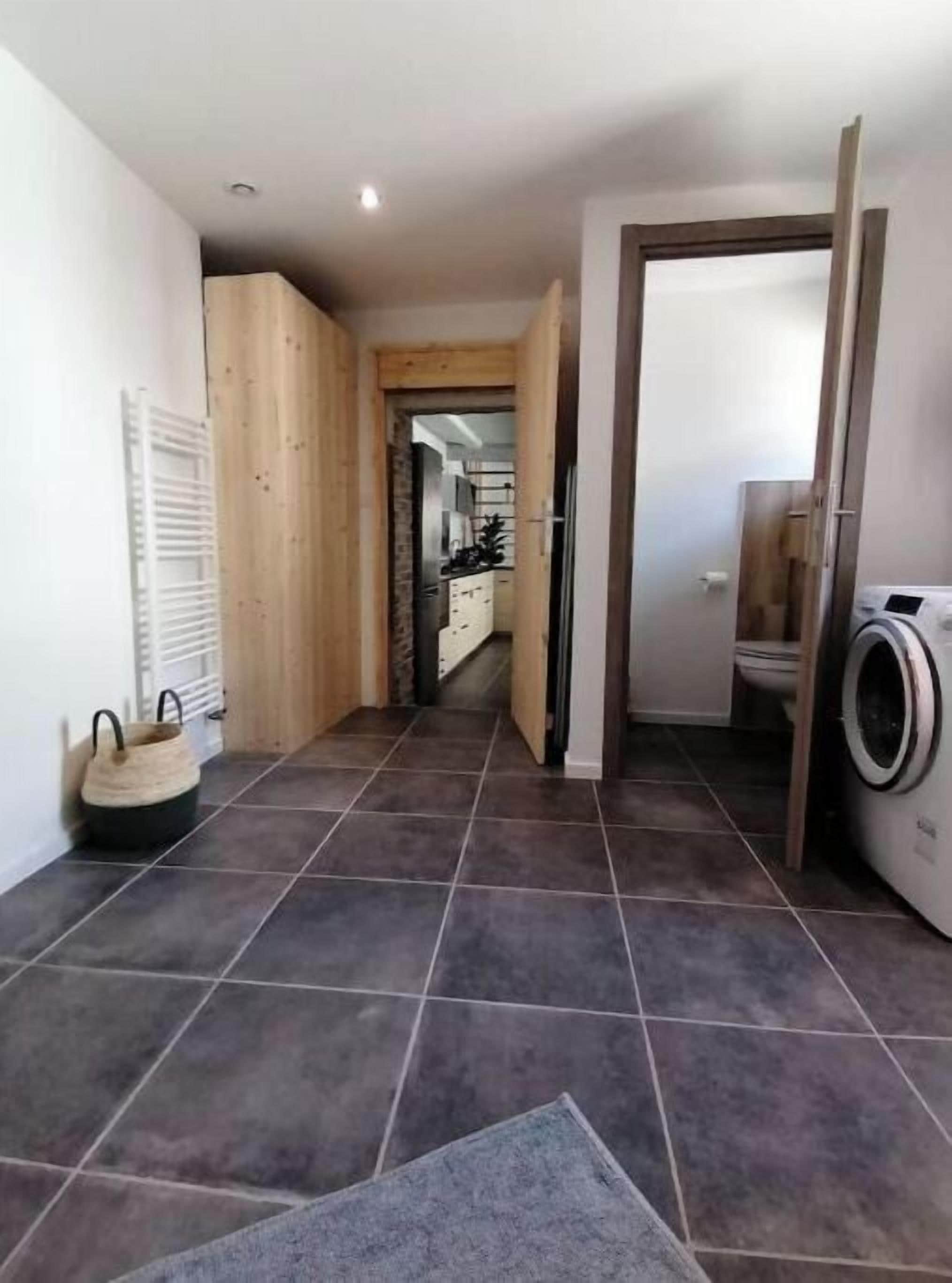 Un couloir avec un sol carrelé, une armoire en bois, un coin buanderie avec un lave-linge, et une cuisine visible au fond.